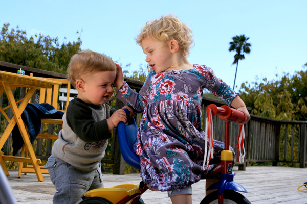 A young girl on a tricycle pushes a young boy away.